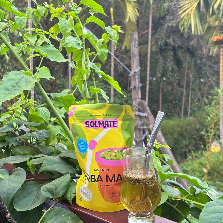 A yellow pouch of Solmaté Yerba Mate  sits on a wooden railing next to a glass of brewed yerba mate tea with a bombilla aka filter straw, surrounded by lush green plants and trees in an outdoor setting